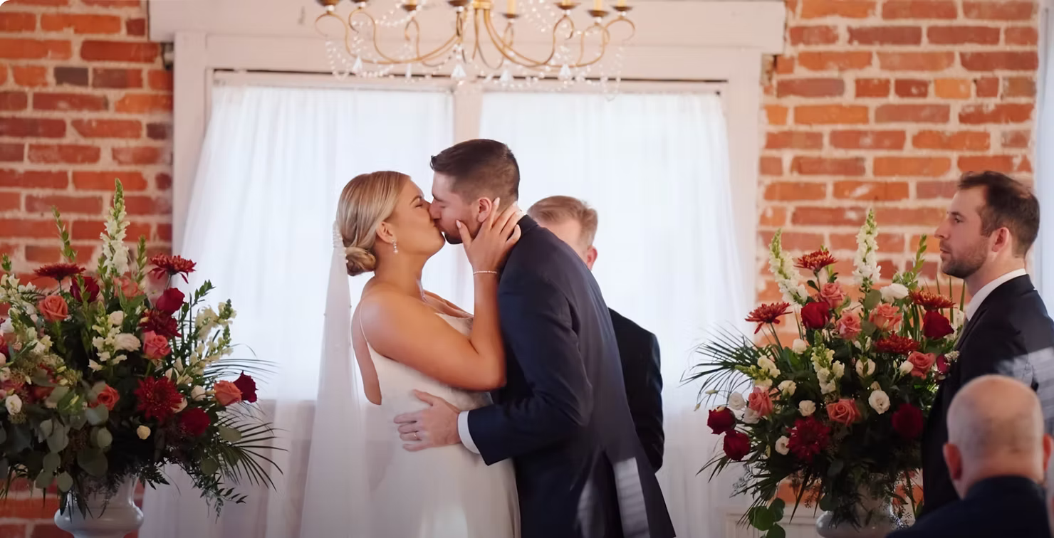 Bride and groom kiss during their ceremony in front of a brick wall with flowers