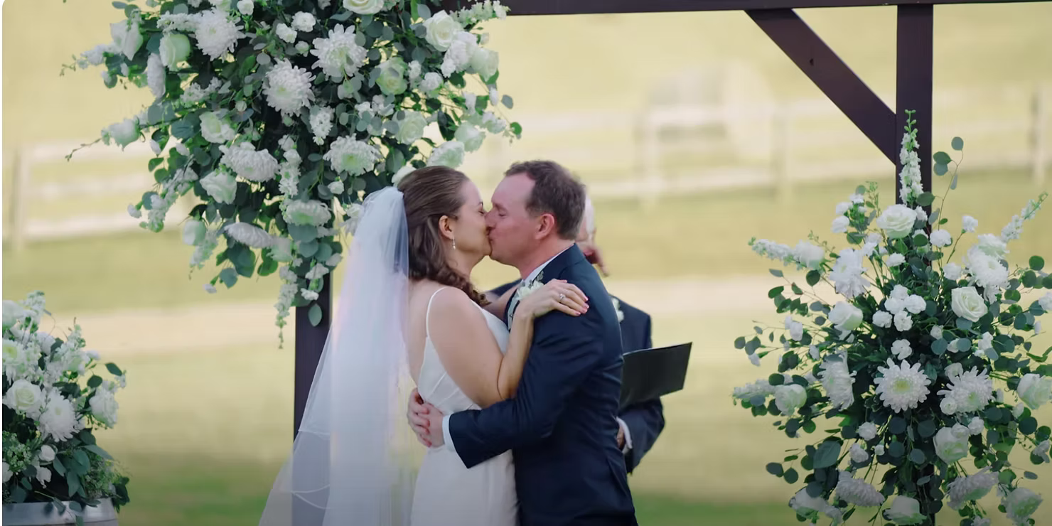 Bride and groom kiss beneath a floral arch at an outdoor ceremony
